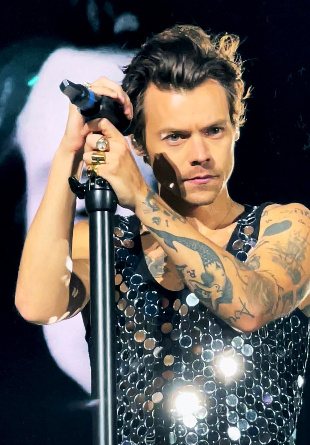 Harry Styles auf der Bühne in einer schwarzen Paillettenweste, Tattoos sichtbar, hält sich am Mikrofonständer fest