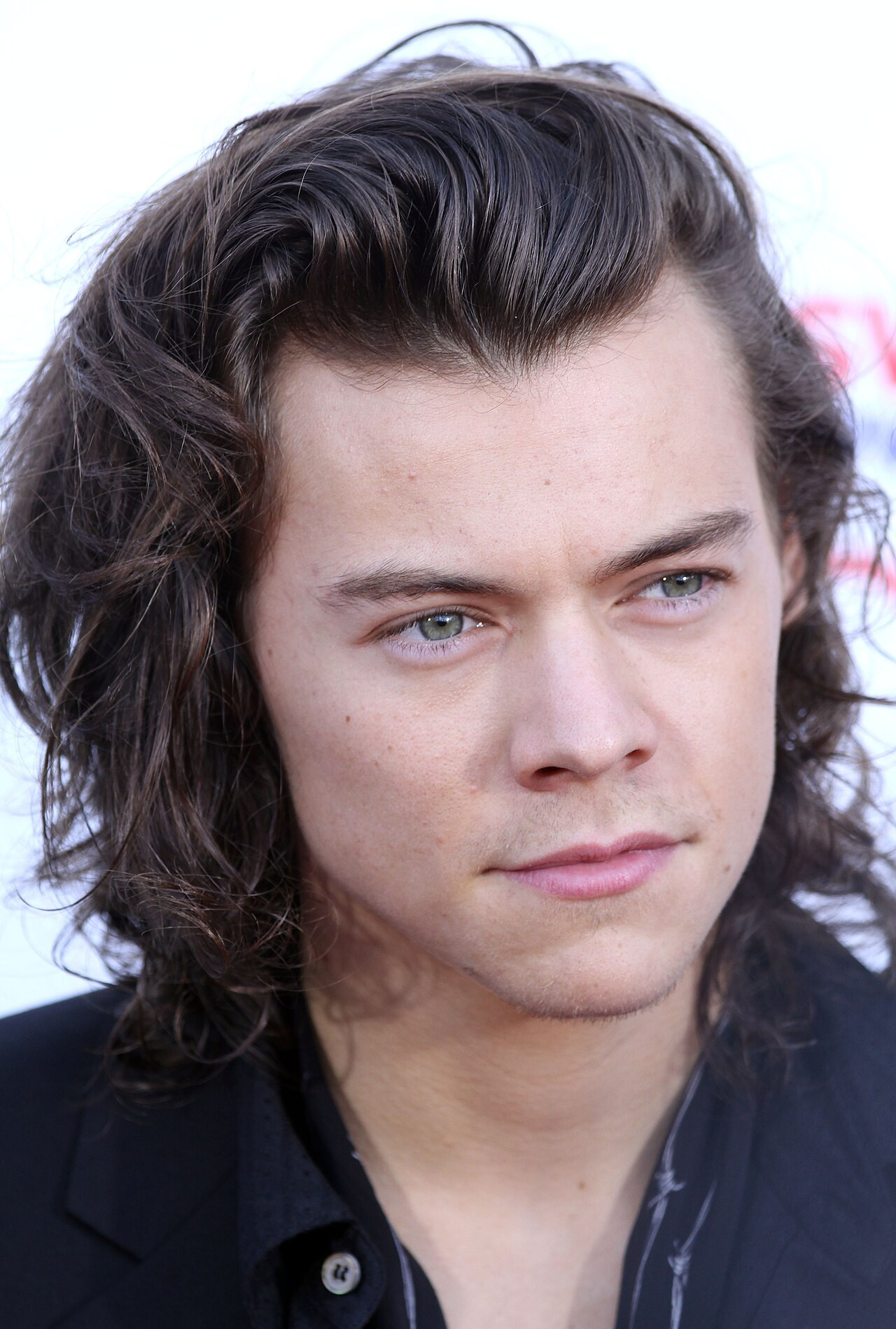 Harry Styles mit langen welligen Haaren in schwarzer Jacke und gestreiftem Hemd auf dem Red Carpet