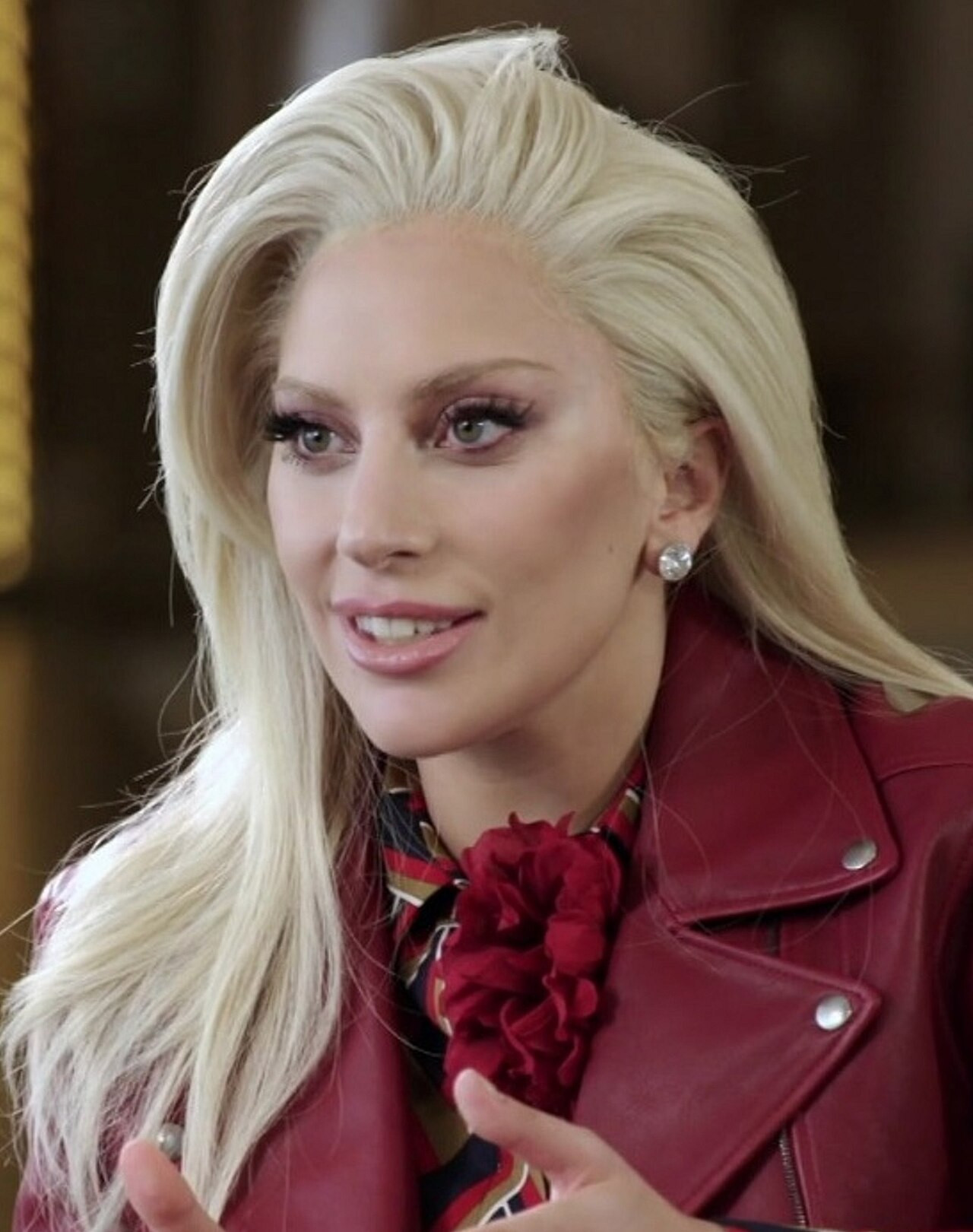 Lady Gaga berambut pirang panjang mengenakan jaket kulit merah dan bros mawar merah.